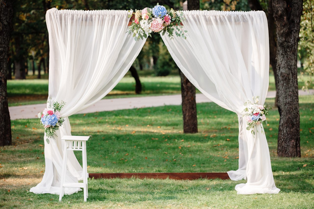Many brides want their wedding ceremony to have a gorgeous and personalized backdrop, because it is in front of this arch that many magic and touching moments occur, and many memorable photos are taken. Wedding arch with flowers and fabrics. Includes: White chiffon fabrics Flowers: white chrysanthemums, pink and light blue hydrangea, assorted greens. Wood stand Dimensions: width 78'', height 86''