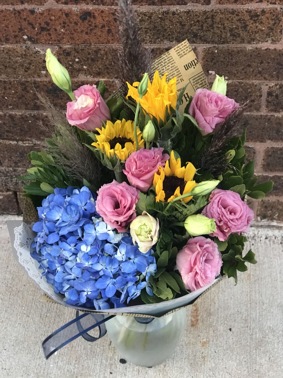 A simply splendid way to surprise someone special! This sophisticated, sunshiny arrangement of hydrangea and roses is an instant pick-me-up!  Includes:  Blue hydrangea, pink lisianthus, sunflowers, assorted greens. Wrapped in a craft paper. Free message card